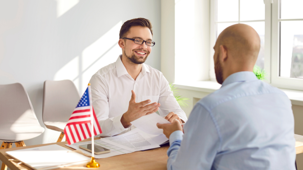 A picture of two people sitting in front of each other and talking about something with a US flag at the table.