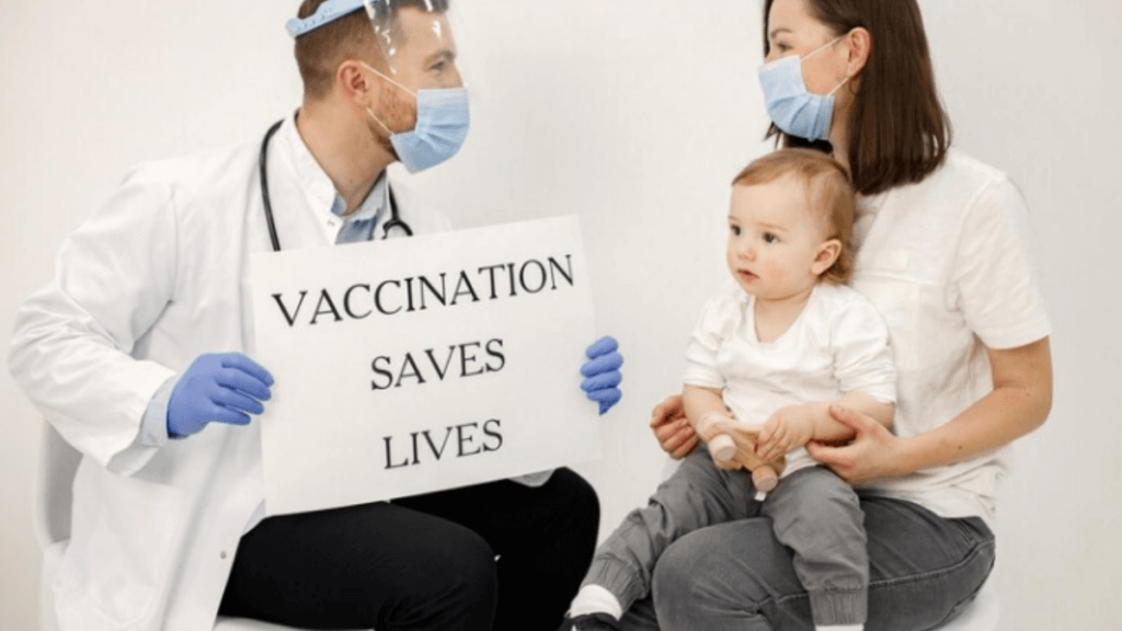 A picture of a doctor and a mother holding her baby in her lap with a cardboard in doctor's hand with "vaccination saves lives" written on it.