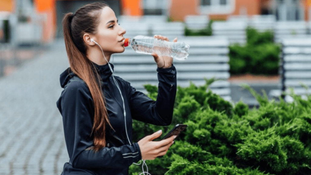 A picture of a girl drinking water with earphones in the ear and holding a mobile phone with a beautiful background.