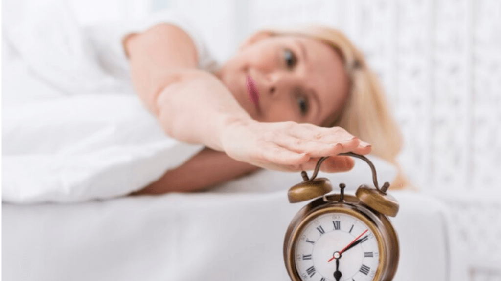 A picture of a lady turning off the alarm clock with lying on a white bed.